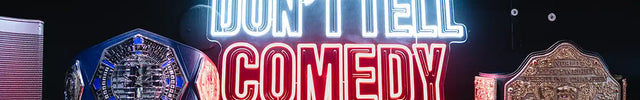 LED neon sign reading 'DON'T TELL COMEDY' in white and red, displayed on a shelf with several championship wrestling belts