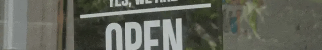 Black acrylic shop sign with white text saying 'Yes, we are open' hanging on a glass door